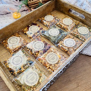 May include: A wooden tray filled with small clear bags containing various dried fruits, herbs, and spices. Each bag has a brown paper label with a handwritten name, such as "Autumn Spice", "Cranberry Orange", and "Lemon & Honey".
