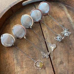 May include: A collection of silver tea infusers with decorative charms, including a bee, teapot, heart, butterfly, and flower. The infusers are arranged on a wooden tray, showcasing their intricate designs and functionality for brewing loose leaf tea.