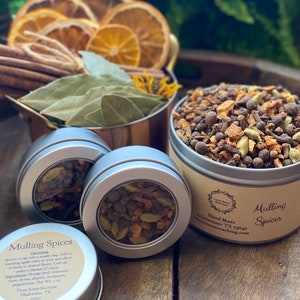 May include: A close-up of three silver tins filled with mulling spices. The tins have clear lids, revealing the spices inside. The spices include cinnamon sticks, cloves, star anise, and orange peel. The label on the largest tin reads "Hand Made Mulling Spices".