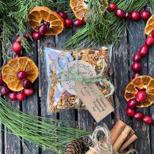 May include: A clear plastic bag filled with a mix of dried fruits, spices, and herbs, tied with twine and a brown paper tag that reads "Holiday Simmering Potpourri Set". The bag is surrounded by dried orange slices, cinnamon sticks, and red berries.