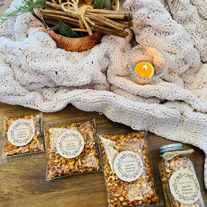 May include: Four bags of apple pie snacking popcorn and a glass jar of apple pie snacking popcorn. The bags and jar have brown paper labels with the text "Apple Pie Snacking Popcorn". The bags and jar are on a wooden surface with a white knitted blanket and a lit candle in the background.