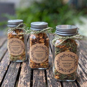 May include: Three glass jars with silver lids and brown paper labels tied with twine. The labels have a wreath design and text that reads "Three Roots Boutique" and the names of the simmering potpourri blends: "Orange Chai", "Sweet Forest", and "Fruit & Orange".