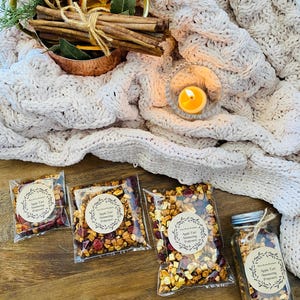 May include: Three clear plastic bags filled with apple tart snacking popcorn, a glass jar filled with the same snacking popcorn, and a copper bowl with cinnamon sticks and dried orange slices. The items are on a wooden surface with a white knitted blanket in the background.