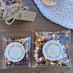 Puede incluir: Dos bolsas transparentes de celof&aacute;n llenas de popurr&iacute; de frutas secas, etiquetadas como "Apple Tart Simmering Potpourri". Una bolsa est&aacute; atada con hilo y una etiqueta que dice "Three Roots Boutique Simmering Potpourri Set". Una vela encendida est&aacute; cerca.
