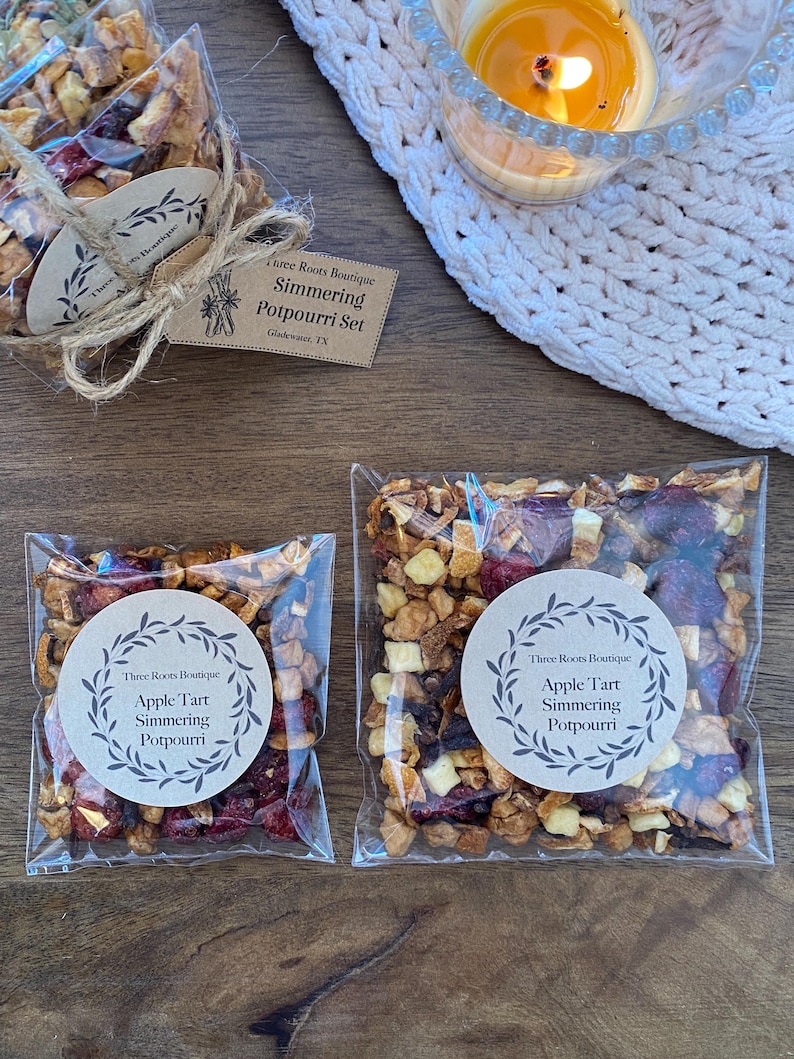 Puede incluir: Dos bolsas transparentes de celof&aacute;n llenas de popurr&iacute; de frutas secas, etiquetadas como "Apple Tart Simmering Potpourri". Una bolsa est&aacute; atada con hilo y una etiqueta que dice "Three Roots Boutique Simmering Potpourri Set". Una vela encendida est&aacute; cerca.