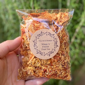 May include: A clear plastic bag filled with dried mango and other fruit pieces for making fruit tea. The bag has a brown label that reads "Three Roots Boutique Mango 'n Friends Fruit Tea".