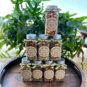 May include: A stack of six glass jars with silver lids and brown labels. The labels have a wreath design and text that reads "Three Room Boutique" and "Orange Chai Simmering Potpourri".
