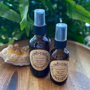 Puede incluir: Dos botellas de vidrio ámbar con pulverizadores y etiquetas de papel marrón. Las etiquetas tienen un diseño de corona y el texto "Three Roots Boutique Light of the Woods Room Spray".