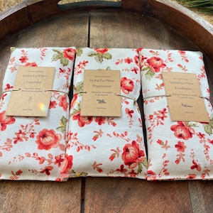 Puede incluir: Tres almohadas para ojos de estampado floral con etiquetas de papel marrón. Las etiquetas dicen "Herbal Eye Pillow Lavender", "Herbal Eye Pillow Peppermint" y "Herbal Eye Pillow Eucalyptus". Cada etiqueta enumera los beneficios del aroma.