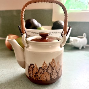 May include: Cream-colored ceramic teapot with a brown lid and a woven handle. The teapot features a forest design in shades of brown. The spout and handle attachments are a light beige color.