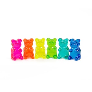 May include: Six translucent gummy bear sweets in a row, in shades of pink, orange, yellow, green, teal, and blue. Each bear has the classic gummy bear shape, set against a white background.