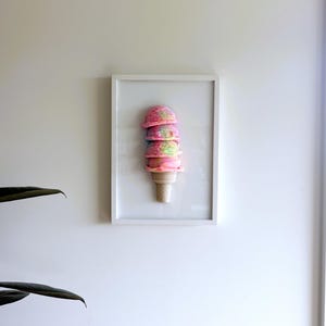 以下が含まれることがあります： 3段のアイスクリームコーンの立体彫刻を収めた額装アート。アイスのスクープはピンク、イエロー、ブルーの鮮やかな色合いがマーブル状に混ざり合い、質感のあるライトベージュのワッフルコーンの上にのっており、白いフレームに収められている。