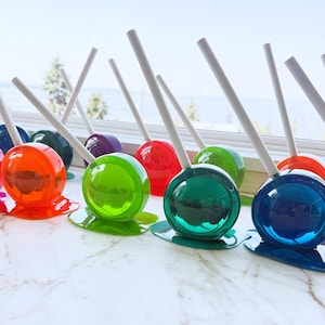 May include: A collection of round, translucent lollipops in various vibrant colors including red, orange, green, blue, and purple. Each lollipop features a smooth, glossy finish with a white stick and a small puddle-like base at the bottom.