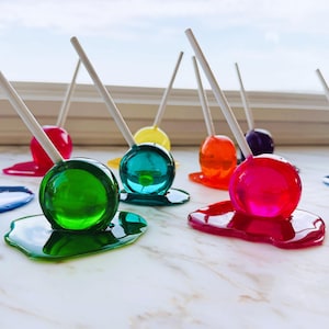 May include: A collection of colorful lollipops on wooden sticks. The lollipops are various colors, including blue, red, green, yellow, orange, pink, and purple. Each lollipop sits on a puddle of matching colored candy.
