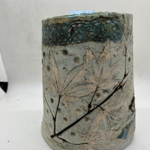 May include: A cylindrical ceramic vase with a textured surface. The vase features a light blue base and body with leaf imprints and dark brown veins. The top section is dark teal with a textured, speckled design. A small, light blue object sits on top.