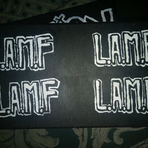 Puede incluir: Tela negra con la palabra "LAMF" repetida cuatro veces en letras blancas. Las letras son estilizadas y tienen un aspecto gráfico audaz.