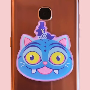 May include: A blue cat-shaped phone grip with large yellow eyes, pink ears, and a pink tongue. The phone accessory has a whimsical design with a small hat on top. The grip is attached to a rose gold phone.