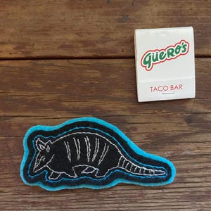 K&ouml;nnte beinhalten: Ein gestickter Armadillo-Aufn&auml;her aus schwarzem Filz mit wei&szlig;en N&auml;hten und einem blauen Umriss. Der Aufn&auml;her befindet sich auf einer Holzoberfl&auml;che.