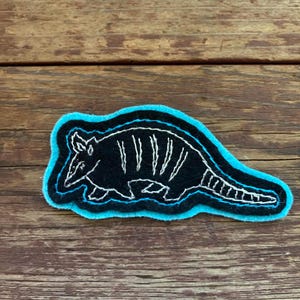May include: A black and white embroidered patch of an armadillo with a blue outline. The patch is on a wooden surface.