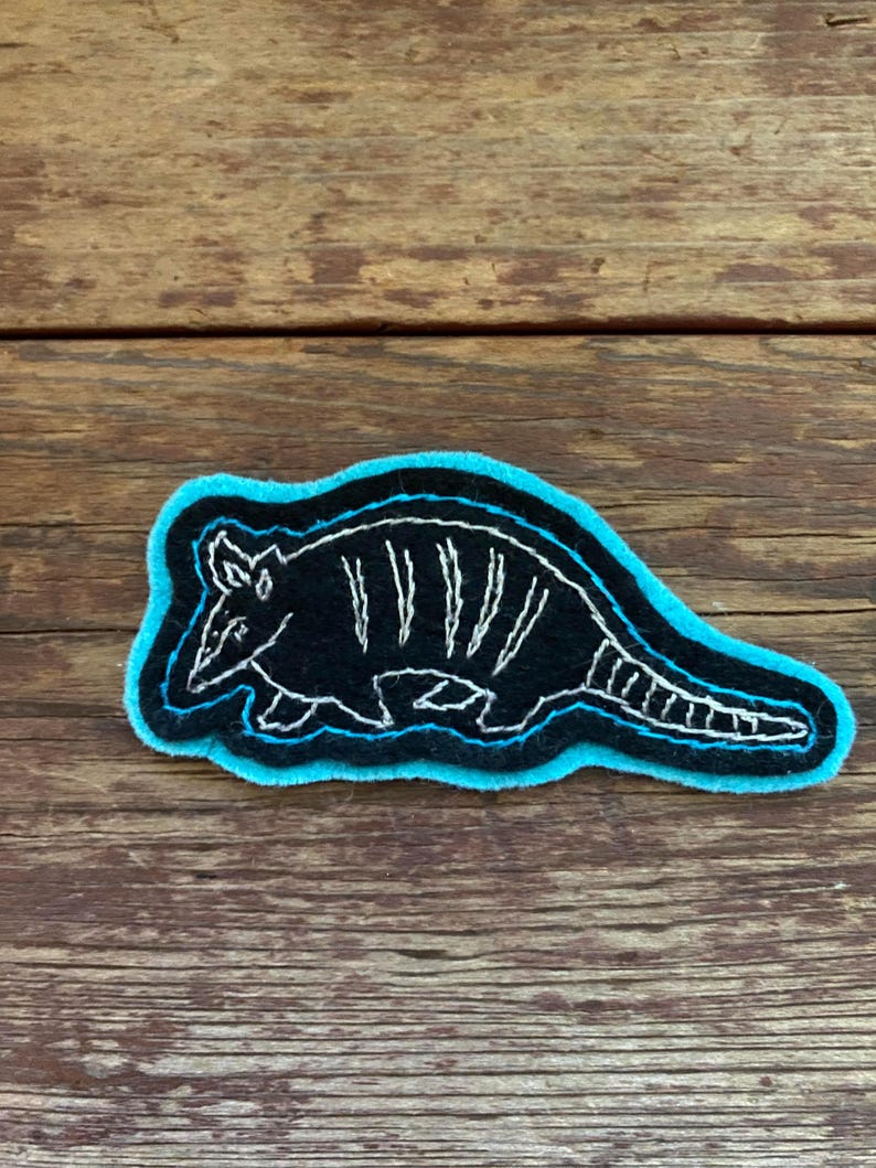 K&ouml;nnte beinhalten: Ein gestickter Patch in Schwarzwei&szlig; mit einem G&uuml;rteltier mit blauem Umriss. Der Patch befindet sich auf einer Holzoberfl&auml;che.
