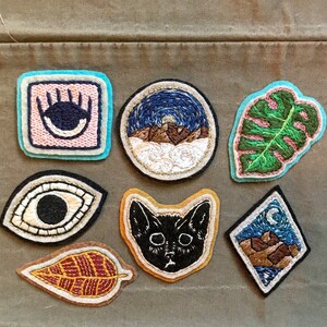 K&ouml;nnte beinhalten: Sieben gestickte Aufn&auml;her mit verschiedenen Designs, darunter eine schwarze Katze, ein Blatt, eine Berglandschaft und ein Auge. Die Aufn&auml;her befinden sich auf einem braunen Stoffhintergrund.