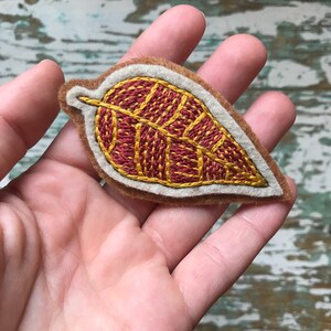K&ouml;nnte beinhalten: Eine Filz-Blatt-Brosche mit einem detaillierten Stickmuster in Rot- und Goldt&ouml;nen.