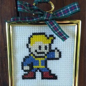 May include: A small framed cross-stitch of a pixelated character from the video game Fallout. The character is wearing a blue jumpsuit and has blonde hair. The frame is gold and has a green and red plaid ribbon attached.