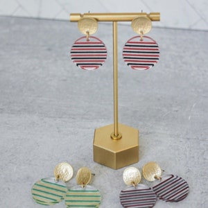 May include: Three pairs of striped earrings with gold accents. The earrings are displayed on a gold stand. The stripes are red and black, green and white, and purple and white.