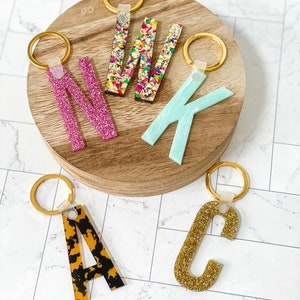 May include: Five colorful letter keychains with gold rings. The letters are N, W, K, A, and C. The letters are made of different materials, including glitter, confetti, and tortoise shell.