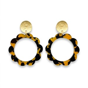 Peut inclure: Une paire de boucles d'oreilles pendantes avec un pendentif circulaire à bords festonnés arborant un motif écaille de tortue noir et jaune. Chaque pendentif est suspendu à un petit disque métallique doré et texturé en haut.