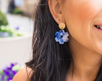 May include: A pair of blue and white floral earrings with a gold disc charm. The earrings are made of resin and have a marbled effect.