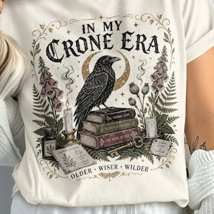 May include: A cream-colored t-shirt featuring a graphic print with the text "IN MY CRONE ERA" at the top and "OLDER WISER WILDER" at the bottom. The design includes a black raven perched on a stack of books, candles, flowers, and a key.