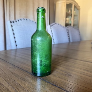 Puede incluir: Una botella de vidrio verde esmeralda de estilo vintage con un cuello delgado y un cuerpo redondeado. El vidrio presenta una superficie ligeramente texturizada y desgastada con signos visibles de antigüedad, incluyendo rasguños menores.