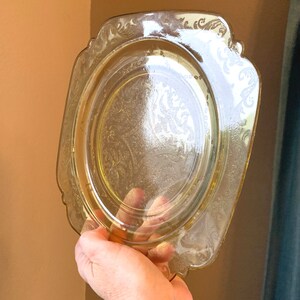 May include: A yellow glass plate with a square shape and a scalloped edge. The plate has a delicate floral pattern etched into the surface.