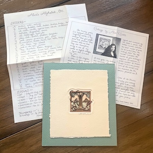 May include: A close-up of a light green card with a cream-colored insert. The insert features a woodblock print of the letter "I" with a butterfly and mountains in the background. The artist's signature is visible at the bottom of the print. The card is titled "Etchings by Alice V. Scott".