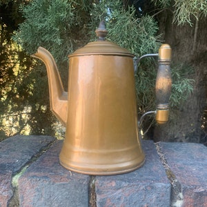 May include: A copper coffee pot with a wooden handle. The pot has a shiny, aged finish and a rounded body.