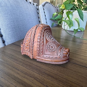 May include: A brown wooden shoe mold featuring intricate hand-carved details, including floral patterns, circular motifs, and geometric borders. The object has a structured, curved shape with a defined heel and toe area, showcasing natural wood grain.