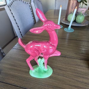 May include: A glossy, bright pink ceramic deer figurine standing on a light green base. The deer is depicted in a leaping pose with one front leg raised, featuring smooth, reflective surfaces and stylized, elongated ears and a slender neck.