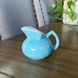 May include: A small, round ceramic creamer pitcher featuring a bright turquoise blue glaze with a speckled, textured finish. The pitcher has a wide pouring spout, a curved handle on the right side, and a smooth white interior surface.