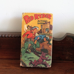May include: A vintage comic book titled "Red Ryder Acting Sheriff." The cover displays a colourful illustration of a cowboy in a red shirt and green trousers, along with other characters in an action scene. The book is set against a yellow background.
