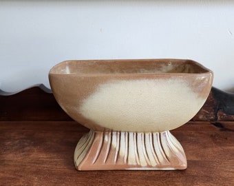 May include: A small, rectangular planter with a brown and cream glaze. The planter has a wide, rounded base and a slightly flared top.