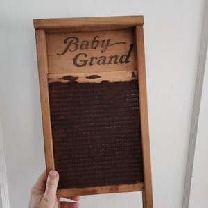 Op de afbeelding: Een vintage houten wasbord met een bruin metalen wasoppervlak. Het wasbord heeft een houten frame en de woorden "Baby Grand" zijn bovenop gedrukt.