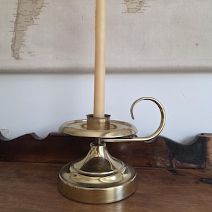 May include: A brass candlestick with a curved handle and a tall, cream-colored candle. The candlestick has a round base and a circular holder for the candle. The background features a vintage world map.