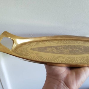 Puede incluir: Una bandeja de servir de color dorado con un diseño adornado. La bandeja tiene un asa en un lado y es de forma ovalada.