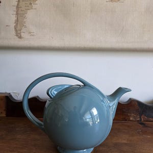 Op de afbeelding: Een teal-kleurige keramische theepot met een afgeronde body, een gebogen handvat en een tuit. De theepot staat op een kleine voet. De achtergrond toont een vintage kaart en een houten oppervlak.