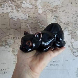 May include: A black ceramic cat figurine with brown eyes. The figurine is being held in a hand against a world map background.