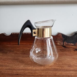 May include: A clear glass coffee pot with a black plastic handle and a gold metal band around the top.