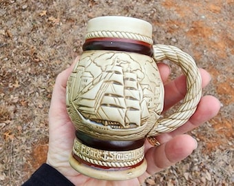 May include: A ceramic beer stein with a rope handle and a detailed image of a sailing ship. The stein has a brown band with the text "The Bark "Staysail" around the bottom.