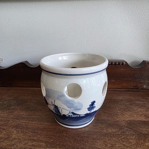 May include: A white ceramic onion keeper with a blue and white landscape design featuring a windmill and trees. The keeper has several round holes and a blue rim. The design is reminiscent of Delftware.
