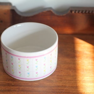 May include: A white ceramic bowl with a pink band and a pattern of colorful tulips. The bowl is cylindrical and sits on a wooden surface. The design includes pink, yellow, blue, and green tulips.